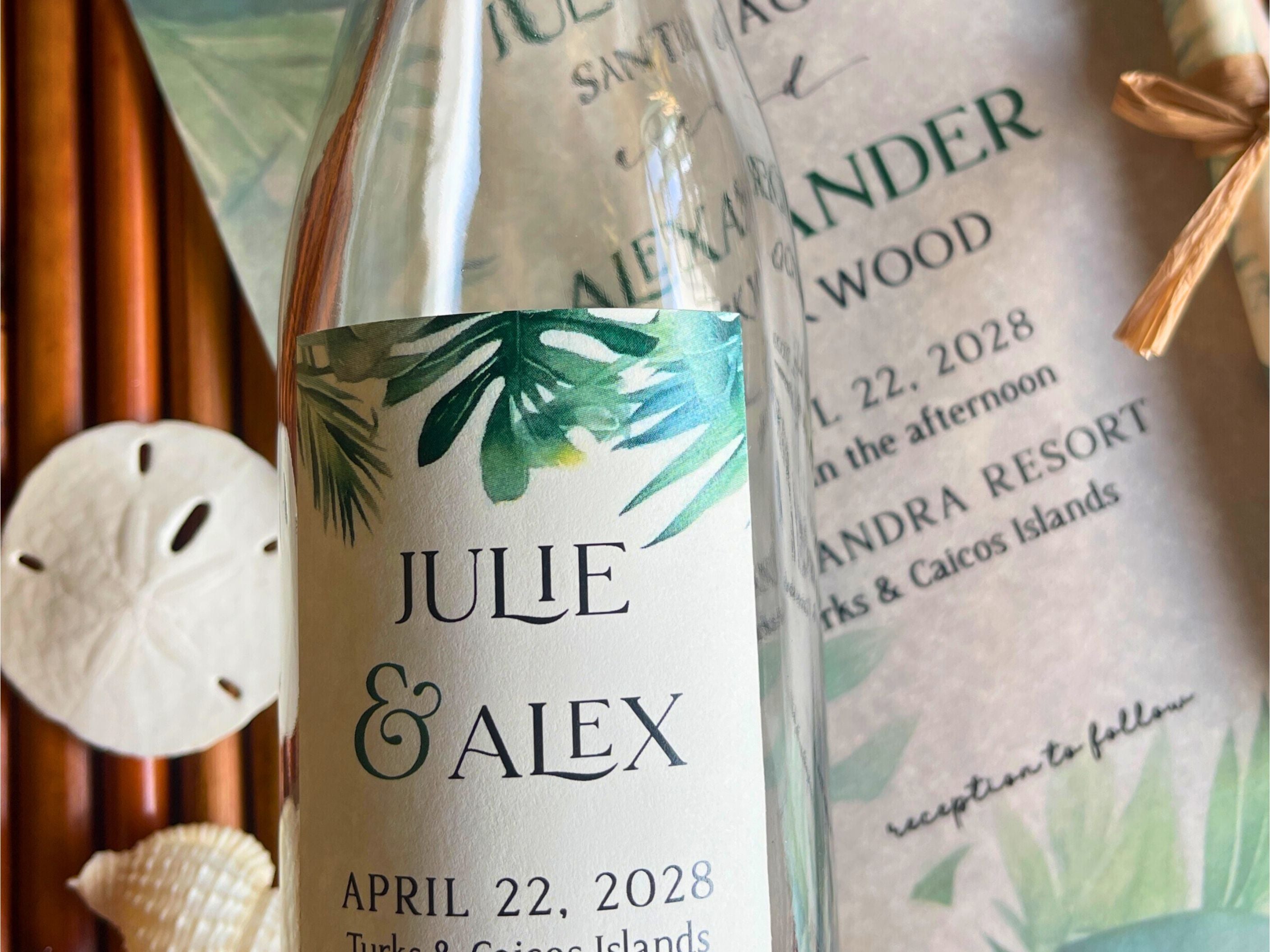 Glass bottle with tropical leaf design label and wedding invitation in the background