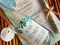 Personalized bottle label with tropical design and wedding details, placed on a woven surface.