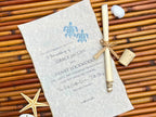 Wedding invitation with blue turtle design on vellum, rolled up with a bow, on a bamboo surface with starfish and shells.