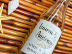 Glass bottle with a personalized label on a woven surface with a shell and starfish.