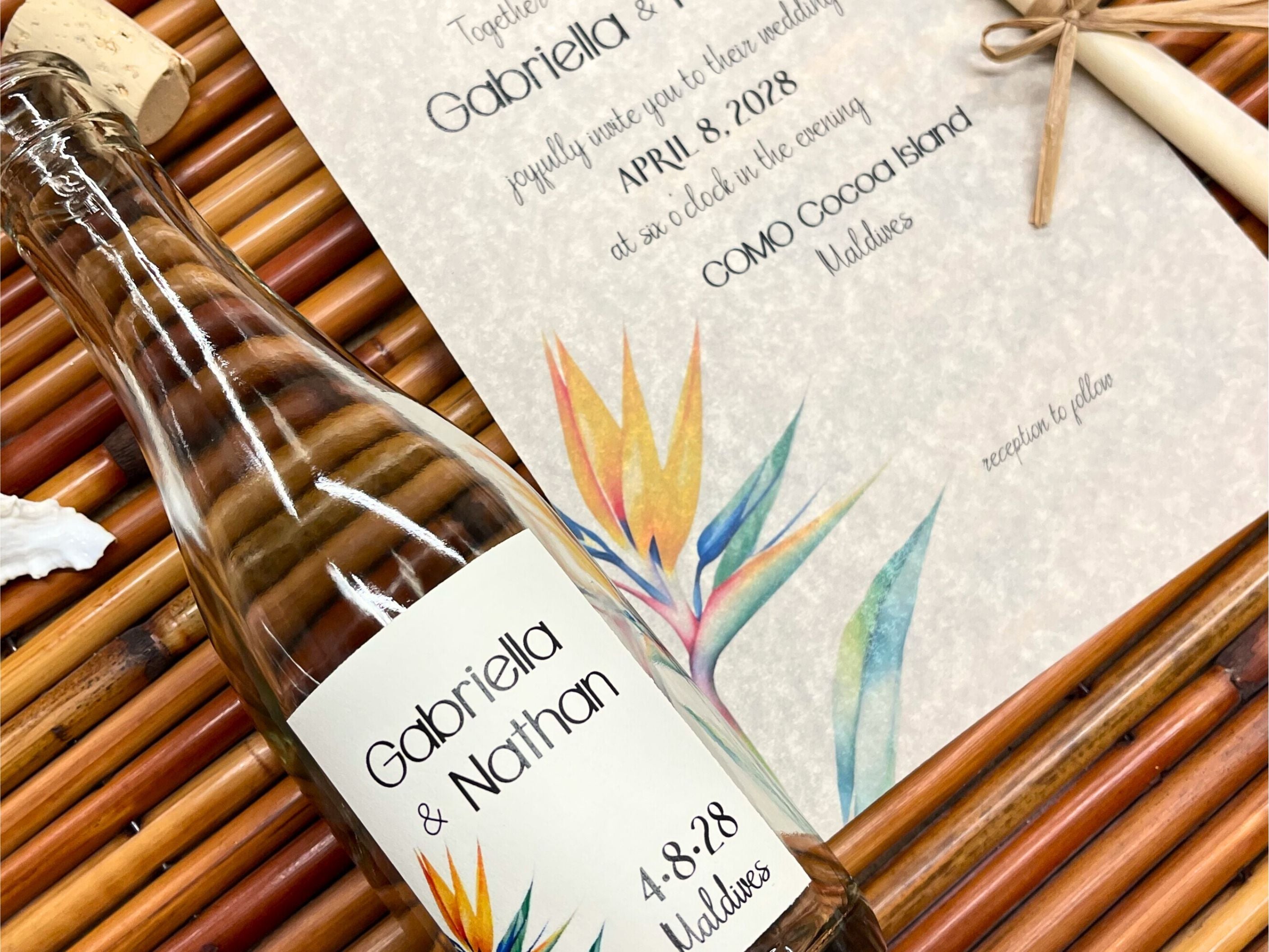 Wedding invitation and glass bottle with a colorful label featuring a tropical bird of paradise plant on a woven mat